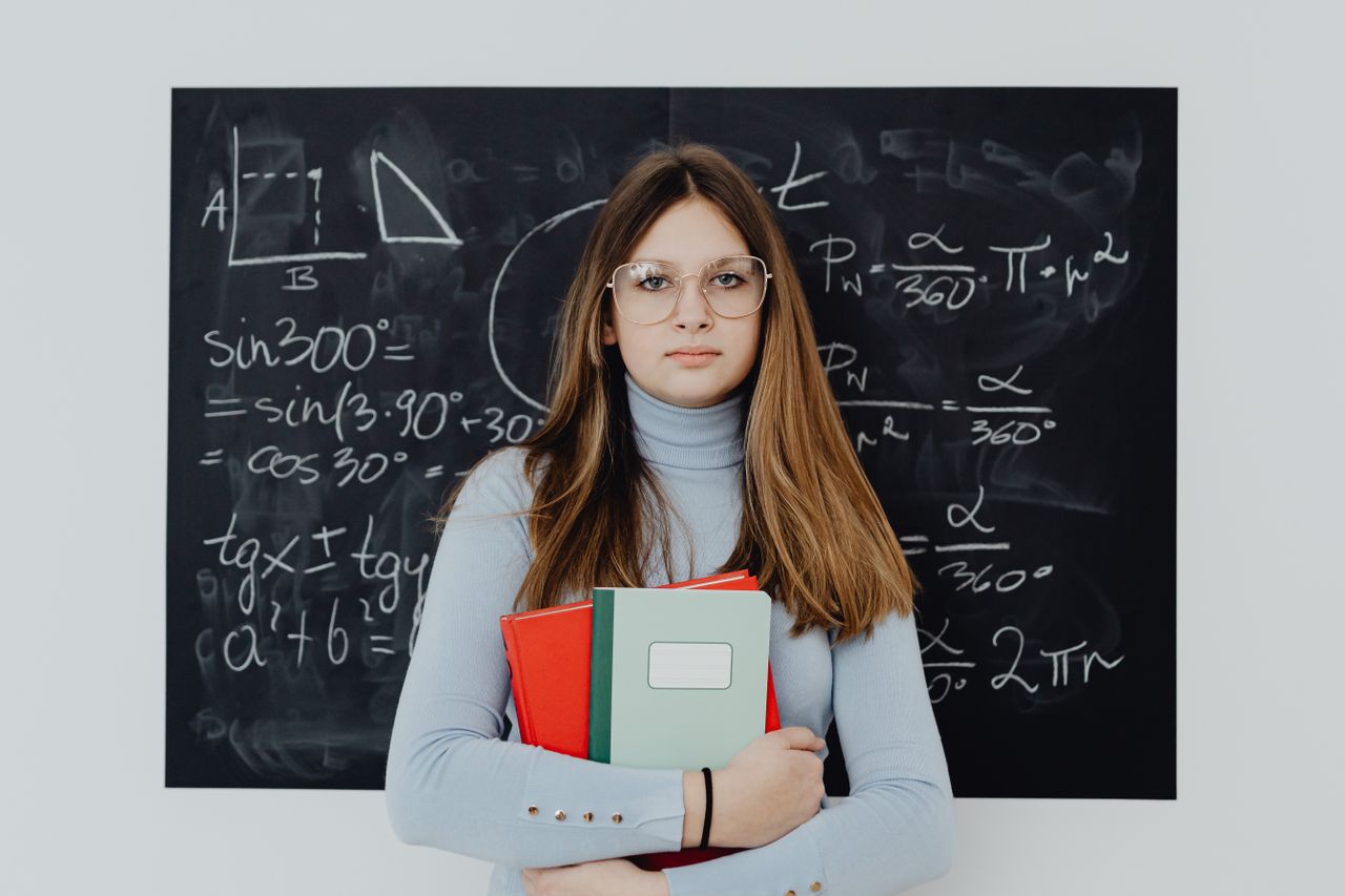 A teenage girl with glasses stands in front of a chalkboard, holding textbooks.