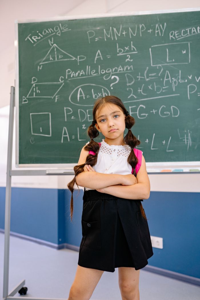 Kendinden emin genç bir öğrenci, matematik, geometrik denklemlerin yazılı olduğu bir kara tahtanın önünde kollarını kavuşturmuş bir şekilde duruyor.